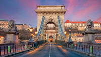 A view of Budapest's historic Chain Bridge at sunset, featuring two stone lion statues on either side of the entrance and the illuminated bridge stretching toward a grand neoclassical archway.
